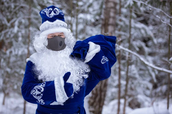 Father Frost in a protective mask with a valve with a bag of gifts in a snowy forest. Social distance, protection against coronavirus. Christmas character of Russia Father Christmas.