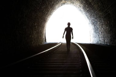 Tünelden çıkan bir kızın silueti. Tünelin sonundaki ışık. Demiryolu rayları ön planda. Taş duvarlar.