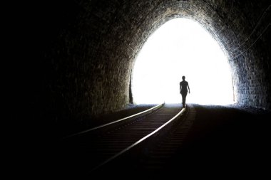 Tünelden çıkan bir kızın silueti. Tünelin sonundaki ışık. Demiryolu rayları ön planda. Taş duvarlar.