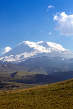 Bir yaz günü karla kaplı dağ zirveleri. Elbrus yeşil tepelerin arkasında. Dağ manzarası. Kafkasya Dağları. Bulutlu mavi gökyüzü. Rusya 'nın doğası.
