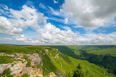 Güneşli bir günde dağ geçidi. Güzel bulutlar, yeşil yamaçlar. Ön planda kayalık bir uçurum. Karachay 'sız cumhuriyet. Rusya
