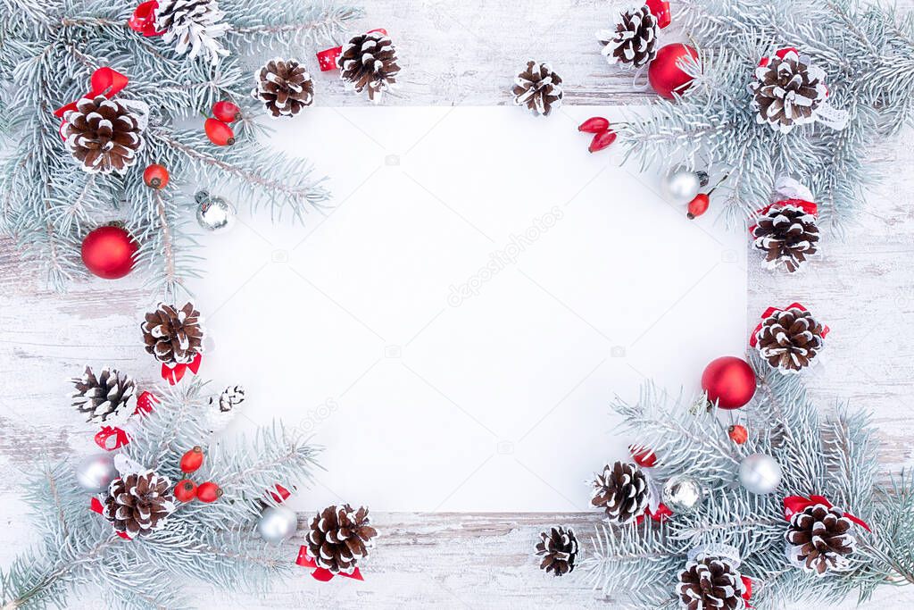 White and red Christmas frame or wreath with blank paper, pine cones and Christmas tree decorations flat lay top view. Stylish Christmas background with snow-covered spruce branches and copy space.