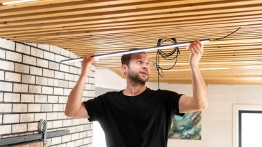 Erkek bir elektrikçi eve elektrik tesisatı kuruyor. Kiracı adam, ahşap tavan kaplamasındaki ışıkları değiştiriyor. Bir inşaatçı tasarımcı tavanına ışık yerleştiriyor.