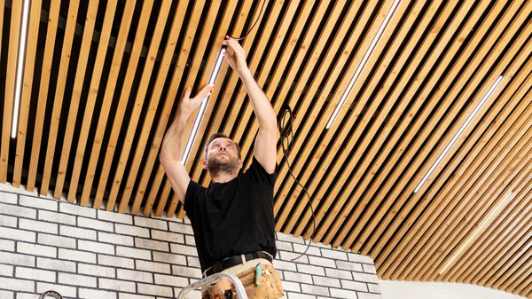 An electrician installs electrical wiring in the house. Installation of a modern ceiling from wooden planks in a loft-style interior. Installing lamps on the ceiling in a new home.