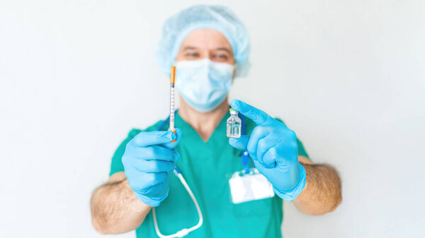 The male doctor smiles and holds a syringe and a vaccine in his hands. Friendly cheerful doctor in disposable cap and mask isolated on white background. Vaccination against COVID-19 during a pandemic.