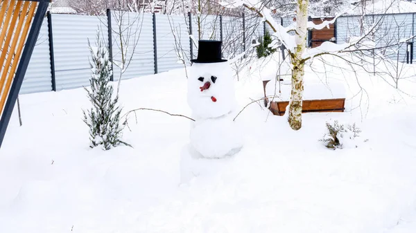 Cute traditional snowman with top hat in a garden swept by snow. Snowman ideas for winter holidays. Winter leisure with children. Christmas garden decorations.