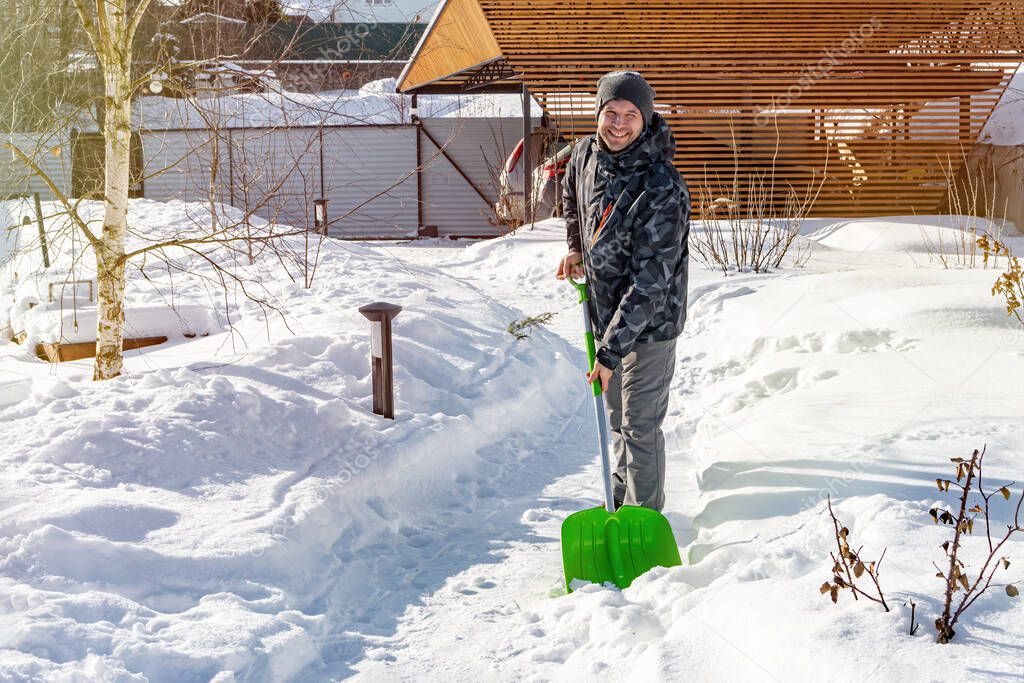 Smiling man cleans the path from snow with a shovel. Cleaning the garden from snow after a heavy snowfall. The yard area is covered with snow and drifts after a snowfall.