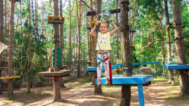 Cesur bir kız halat kasabasındaki ağaçlar arasında uzanan ince bir ip boyunca yürüyor. Çocuklar için aktif spor eğlencesi. Çocuklarda cesaret ve fiziksel dayanıklılık geliştirmek için açık hava eğitimi.