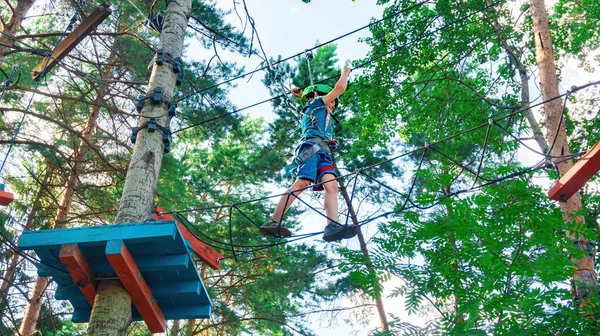 A brave boy walks along a rope ladder stretched between trees at a great height. Family fun in the adventure high rope park. Extreme kinds of entertainment for children during summer holidays.
