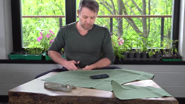 Мужчина режет ткань для шитья фартука к зеленым деревьям в окне. — стоковое видео