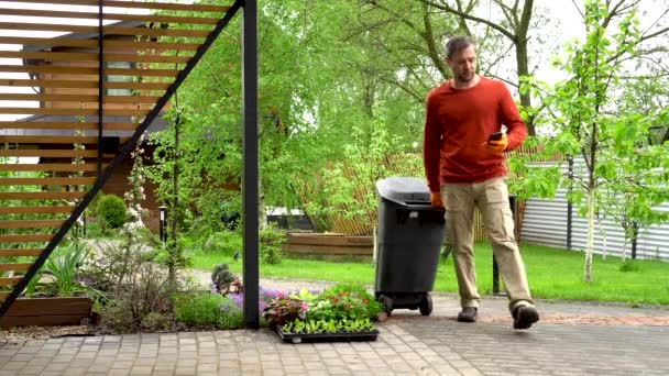 Een huiseigenaar kijkt naar zijn telefoon als hij een vuilnisbak buiten zet.. — Stockvideo