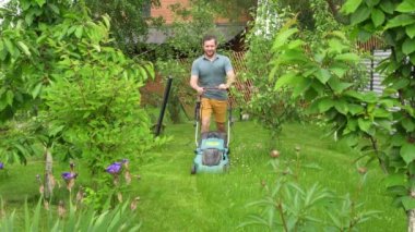 Genç meyve ağaçları arasında elektrikli çim biçme makinesiyle çimleri biçen bir adam.. 