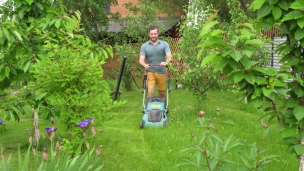 En man klipper gräset med en elektrisk gräsklippare mellan unga fruktträd. — Stockvideo