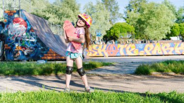 Portrait of a smiling girl with a skateboard in her hands. A cute girl in a helmet and knee pads spends time in the extreme sports park. The concept of sports in the life of teenagers.