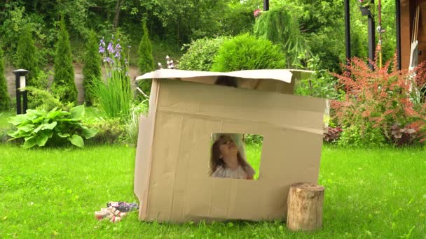 Flickor leker i ett kartonghus nära huset på gräsmattan. — Stockvideo