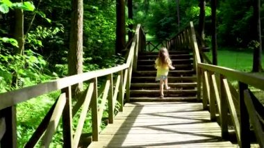 Bir kız ağaçların arasındaki parkta tahta bir güvertede koşuyor.. 