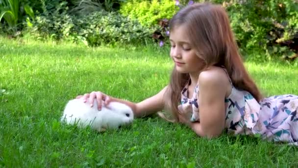 Ένα κορίτσι χαϊδεύει ένα χαριτωμένο λευκό κουνέλι στο γρασίδι κοντά στο σπίτι σε μια ηλιόλουστη καλοκαιρινή μέρα. Το παιδί παίζει με ένα κατοικίδιο. Στο φόντο ενός παρτέρι με λουλούδια. — Αρχείο Βίντεο