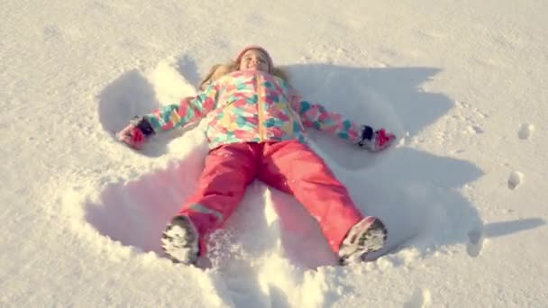 En liten söt flicka i ljusa vinterkläder gör en snöängel i snön. — Stockvideo