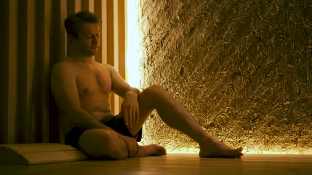 A handsome man with a naked torso is siting on a wooden floor in a sauna. — Stock Video