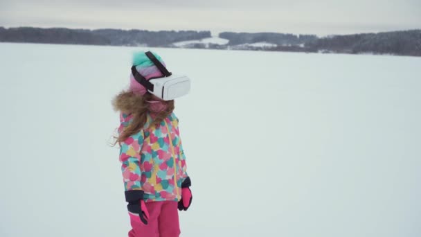 En flicka i ljusa kläder och virtuella verklighets glasögon står på en snö. — Stockvideo