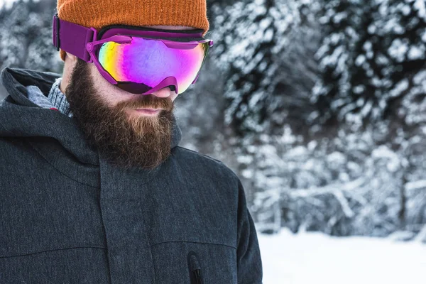 Um homem adulto brutal com uma barba em óculos de esqui e um chapéu fica nas montanhas no inverno. Conceito de recreação ativa — Fotografia de Stock