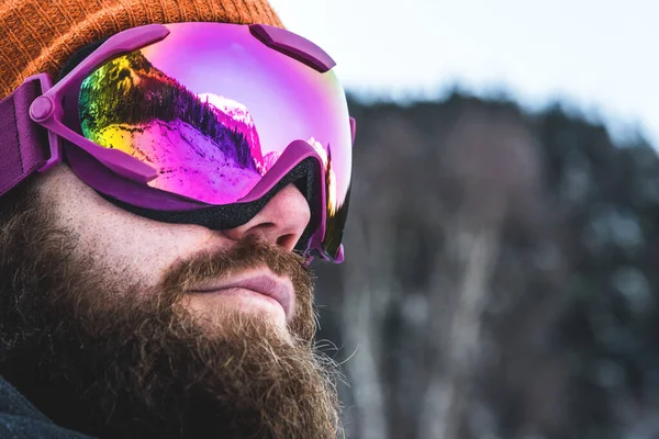 Een volwassen brutale man met een baard in skibril en een hoed staat in de winter in de bergen. Actief recreatieconcept — Stockfoto