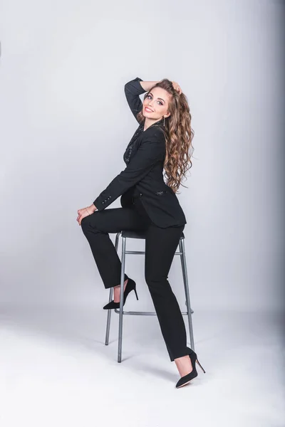 Uma mulher bonita em um terno de negócios preto com uma maquilagem e cabelo longo senta-se em uma cadeira alta em um fundo branco no estúdio. Conceito de mulher de trabalho e negócios — Fotografia de Stock