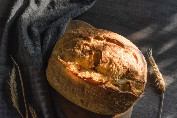 Ręcznie robiony tartinowy chleb leży na drewnianej powierzchni obok kłosów pszenicy. Koncepcja produkcji rolnej i pieczenia w domu — Zdjęcie stockowe