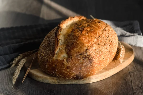 Ręcznie robiony tartinowy chleb leży na drewnianej powierzchni obok kłosów pszenicy. Koncepcja produkcji rolnej i pieczenia w domu — Zdjęcie stockowe