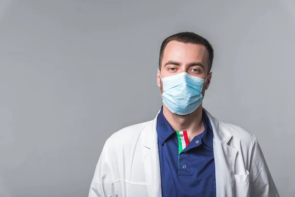 Um jovem com roupas médicas em uma máscara e luvas fica em um fundo cinza no estúdio. Conceito de cuidados de saúde e serviço médico — Fotografia de Stock