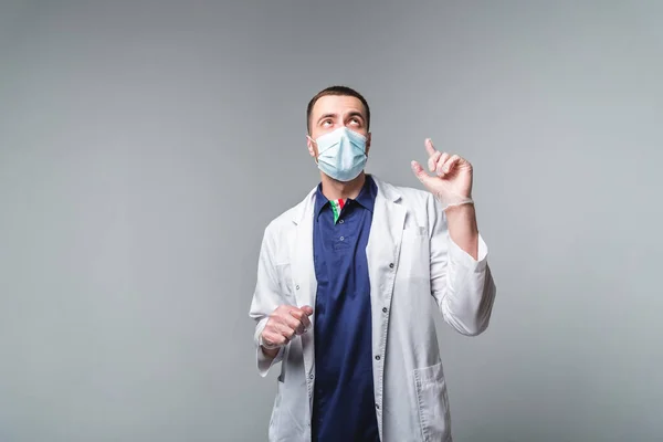 Молодой человек в медицинской одежде в маске и перчатках стоит на сером фоне в студии. Здравоохранение и медицинское обслуживание — стоковое фото