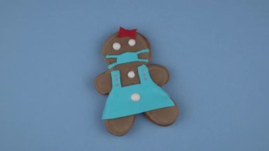 Mavi arka planda tıbbi maske takmış zencefilli kek. Tıbbi solunum bandajı, cerrahi koruyucu maske. Kurabiyeler el yapımı. Döndürme hareketi. Pandemik Coronavirus sırasında Noel