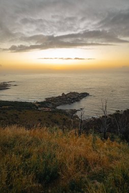 Sahil manzarası, sakin okyanusa bakan kuru otlar ve gün batımında kayalık kıyılar. Seyahat ve çevre kavramı için doğal arka plan.