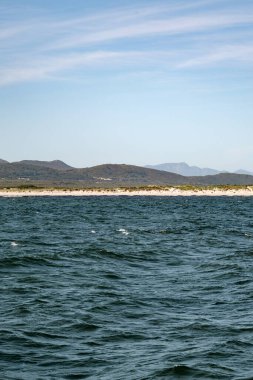 Dalgalı koyu mavi okyanus, açık mavi gökyüzünün altındaki uzak yeşil tepeler. Seyahat ve doğa konsepti için doğal deniz manzarası.