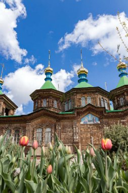 Yeşil çatıları ve mavi gökyüzünün altında beyaz bulutlu altın kubbeleri olan, ön planda laleler bulunan eski ahşap bir kilise. İbadet için dini mimari.