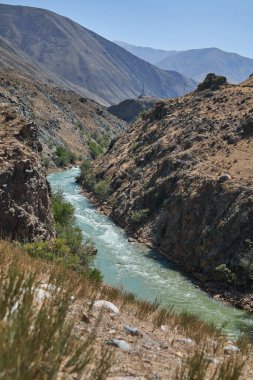 Dağ nehri, engebeli doğal bir manzarayı tasvir eden açık mavi gökyüzünün altında sarp kayalık yamaçlarla derin bir kanyon vadisinde akıyor..