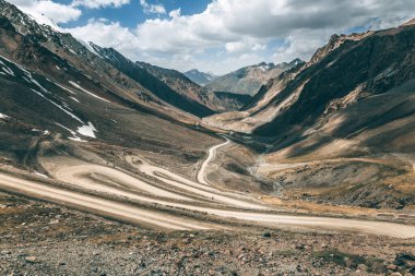 Bulutlu bir gökyüzünün altında kayalar ve karla kaplı tepeler olan bir dağ geçidinden geçen toprak bir yol. Macera yolculuğu konsepti için engebeli arazide yolculuk.