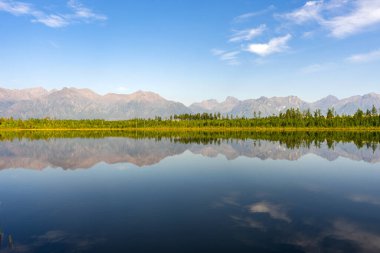 Dağları ve ormanı olan sakin bir göl mavi gökyüzünün altındaki suya yansıyor. Seyahat broşürü için doğal manzara, doğa geçmişi..