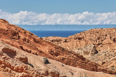 Açık gökyüzü ve beyaz bulutlarla mavi bir göle uzanan kızıl dağ kayalarından oluşan güzel bir manzara. Tien Shan Dağları 'na seyahat edin..