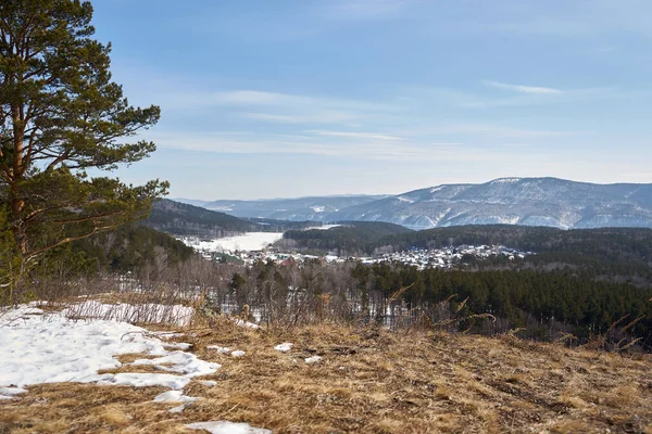 Kuru çimler ve kısmi karla kaplı tepe manzarası. Kış köyü, yemyeşil orman ve doğa yolculuğu için mavi kış gökyüzü altındaki dağlar..