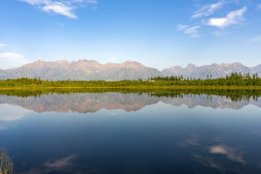 Mavi gökyüzünün altındaki orman ve dağların yansımalarıyla sakin göl seyahat ve tatil için doğadaki huzuru temsil ediyor. Tayozhnoye Gölü Chara kumları yakınında, Kodar sıradağları.