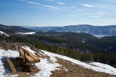 Arka planda orman ve kasaba olan karlı bir dağda boş bir bank. Seyahat ve doğa keşfi için kış manzarası.