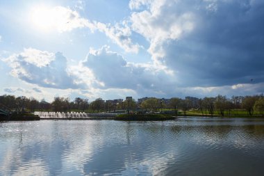 Parçalı bulutlu bir gökyüzünün altındaki şehir parkındaki fıskiyeli büyük bir göletin manzarası, gökyüzünü ve etrafındaki ağaçları yansıtan sakin bir su yüzeyini gösteriyor..