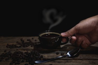 Bir adamın eli, eski bir ahşap masada kahve fincanını siyah bir fonda tutuyor..