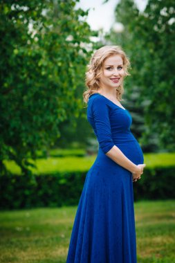 Beautiful young pregnant woman in blue dress with long blond curly hair holding her belly and looking at camera in summer park on rainy day. Pregnancy and femininity concept. Waterdrops on blue dress
