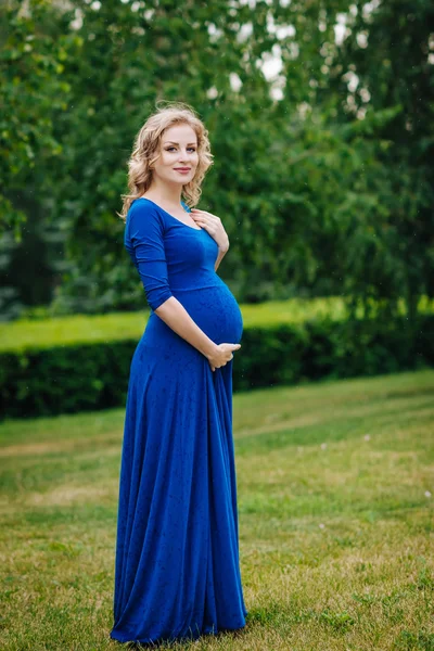 Mooie jonge zwangere vrouw in blauwe jurk met lang blond krullend haar haar buik in zomerpark op regenachtige dag houden. Zwangerschap en vrouwelijkheid concept. Waterdruppels op blauwe jurk — Stockfoto