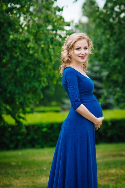 Schöne junge schwangere Frau in blauem Kleid mit langen blonden lockigen Haaren, die ihren Bauch hält und an einem regnerischen Tag im Sommerpark in die Kamera schaut. Schwangerschaft und Weiblichkeit. Wassertropfen auf blauem Kleid — Stockfoto