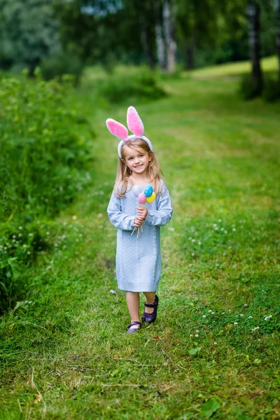 Ragazzina sorridente con lunghi capelli biondi che indossa orecchie di coniglio o coniglio e abito blu a maglia. Carina bambina che tiene un mucchio di uova colorate e cammina nel parco primaverile. Celebrazioni pasquali — Foto Stock