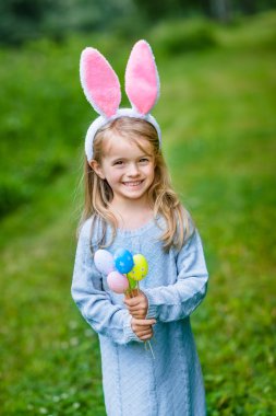 Portrait of smiling little girl with long blond hair wearing rabbit or bunny ears and blue knitted dress and holding bunch of painted colour eggs on sunny day in spring park. Easter celebrations