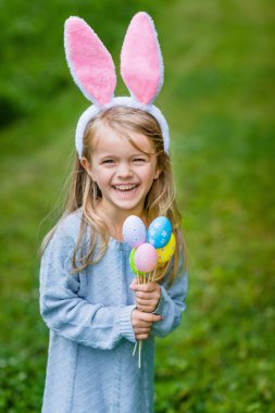 Adorable laughing little girl with long blond hair wearing pink rabbit or bunny ears and blue knitted dress and holding bunch of painted colour eggs on sunny day in spring park. Easter celebrations